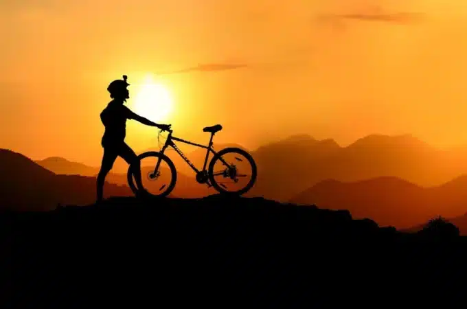 Ciclista de mountain bike em silhueta contra um pôr do sol alaranjado, com montanhas ao fundo, simbolizando a aventura do MTB.