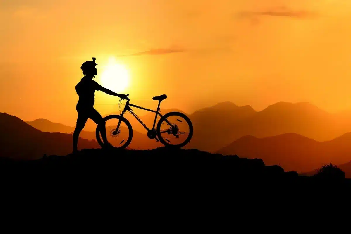 Ciclista de mountain bike em silhueta contra um pôr do sol alaranjado, com montanhas ao fundo, simbolizando a aventura do MTB.