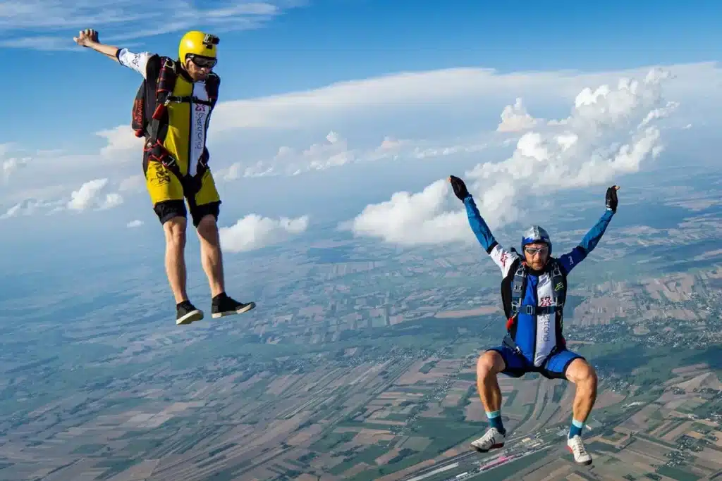 Dois paraquedistas em queda livre, flutuando acima de uma vasta paisagem verde de campos e nuvens brancas, demonstrando a emoção das altitudes elevadas.