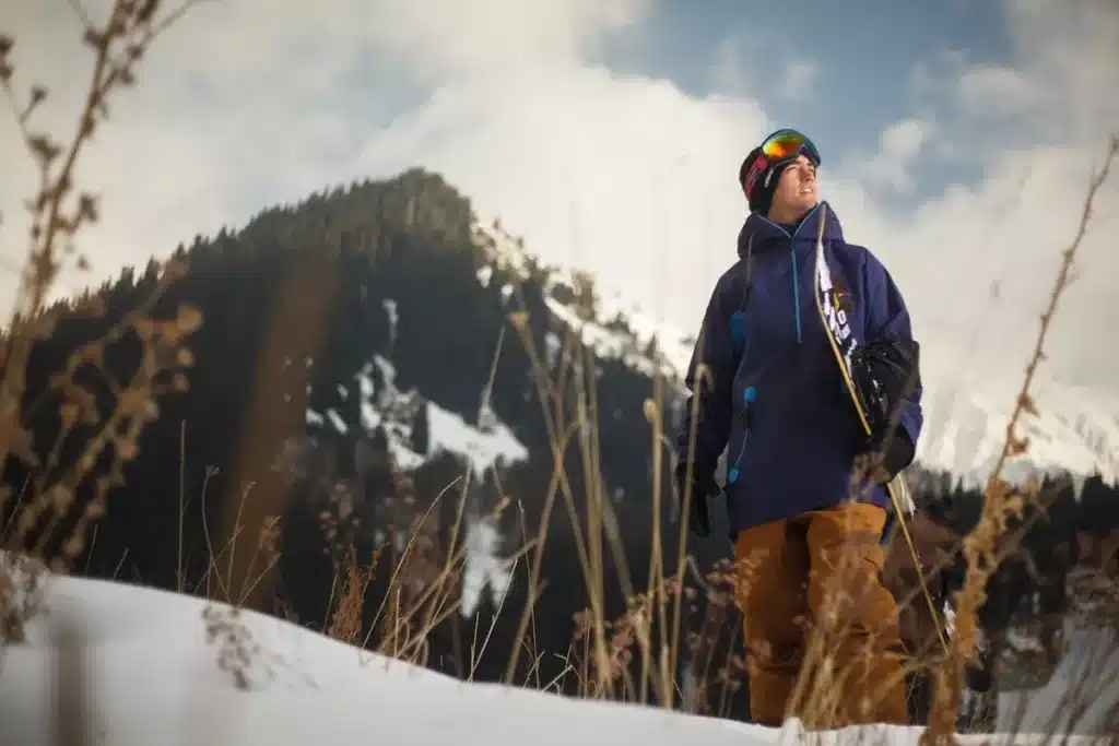 Snowboarder com óculos de proteção e jaqueta azul-marinho, segurando sua prancha e olhando para o horizonte em uma paisagem de neve com montanhas e vegetação seca em altitudes elevadas.