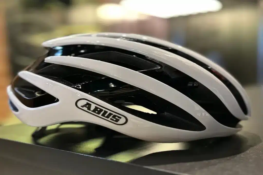 Um capacete de mountain bike masculino branco e preto da marca ABUS, com design aerodinâmico e aberturas de ventilação, posicionado sobre uma superfície escura, pronto para uso em trilhas.