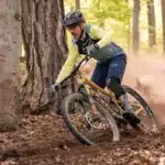 Um ciclista masculino em alta velocidade usando um "capacete mountain bike masculino" escuro, óculos de proteção, camisa de manga longa amarela e azul, e shorts acolchoados, manobrando sua bicicleta em uma trilha de terra com árvores ao redor, levantando poeira.