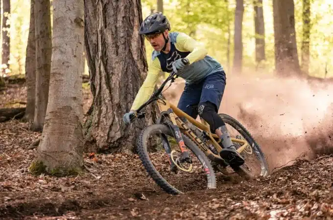 Um ciclista masculino em alta velocidade usando um "capacete mountain bike masculino" escuro, óculos de proteção, camisa de manga longa amarela e azul, e shorts acolchoados, manobrando sua bicicleta em uma trilha de terra com árvores ao redor, levantando poeira.