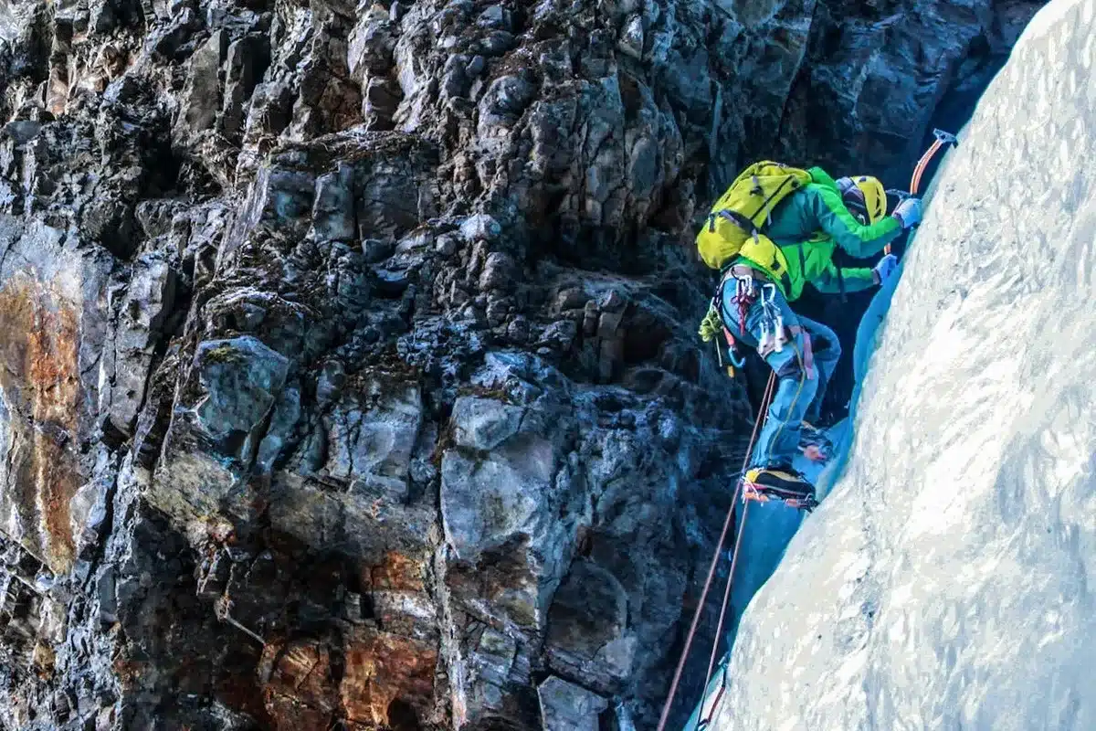 Alpinista equipado com picadores de gelo e mochila amarela, escalando uma parede íngreme de gelo ao lado de uma rocha em altas altitudes.