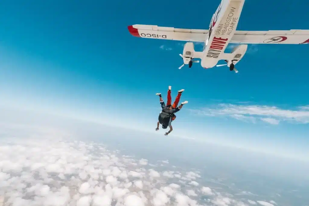 Dupla realizando salto de paraquedas em queda livre, com o avião ao fundo e céu azul sobre nuvens.