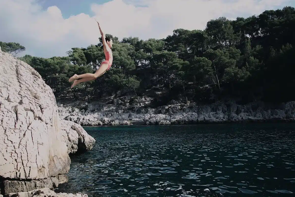 Mulher em queda livre ao saltar de um penhasco para o mar, em meio à natureza e águas cristalinas.