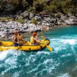 Quatro pessoas vestindo coletes salva-vidas e capacetes remando em bote inflável amarelo sobre corredeiras de rio turquesa, com margens rochosas e vegetação ao fundo