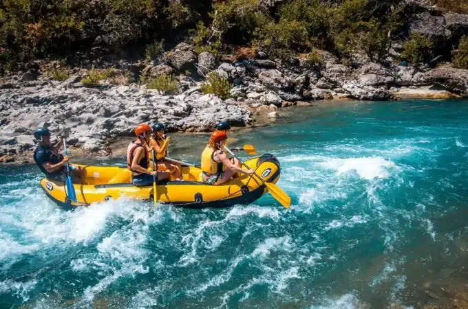 Quatro pessoas vestindo coletes salva-vidas e capacetes remando em bote inflável amarelo sobre corredeiras de rio turquesa, com margens rochosas e vegetação ao fundo