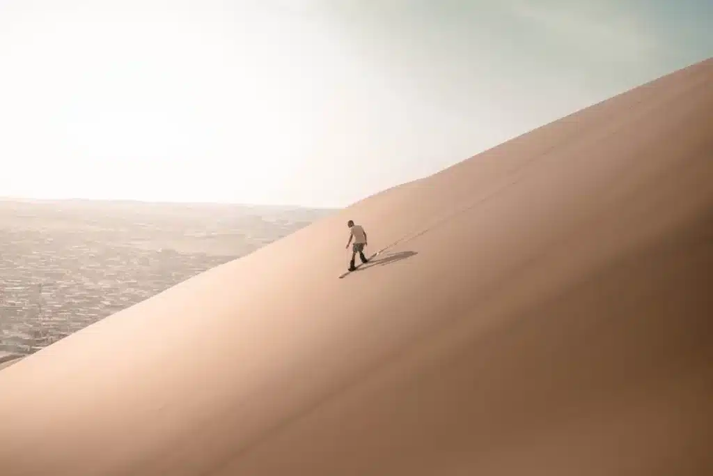 Uma pessoa solitária vestindo camiseta branca e shorts escuros pratica sandboard em uma vasta e alta duna de areia dourada sob um céu claro com leve neblina. Uma cidade pode ser vista ao longe, na base da duna.
