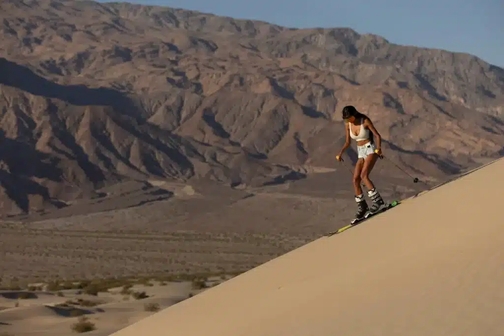 Uma mulher negra em trajes de esqui brancos e shorts jeans desce uma duna de areia clara com equipamentos de esqui em um dia ensolarado, com montanhas áridas ao fundo.