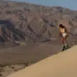 Uma mulher negra em trajes de esqui brancos e shorts jeans desce uma duna de areia clara com equipamentos de esqui em um dia ensolarado, com montanhas áridas ao fundo.