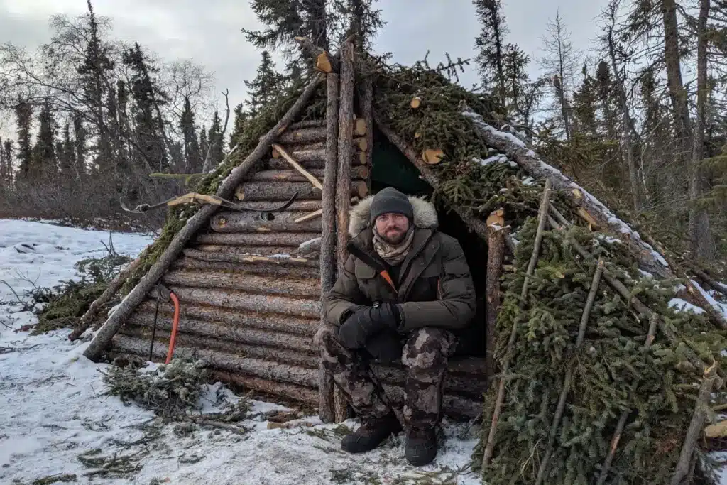 Um homem barbudo, vestido com um casaco de inverno verde-oliva e roupas camufladas, está sentado na entrada de um abrigo tipo lean-to ou cabana de madeira e galhos construída por ele mesmo, coberta de neve e pinhas. A cena, em uma floresta nevada, representa a arte do Bushcraft e as técnicas de sobrevivência no frio extremo.