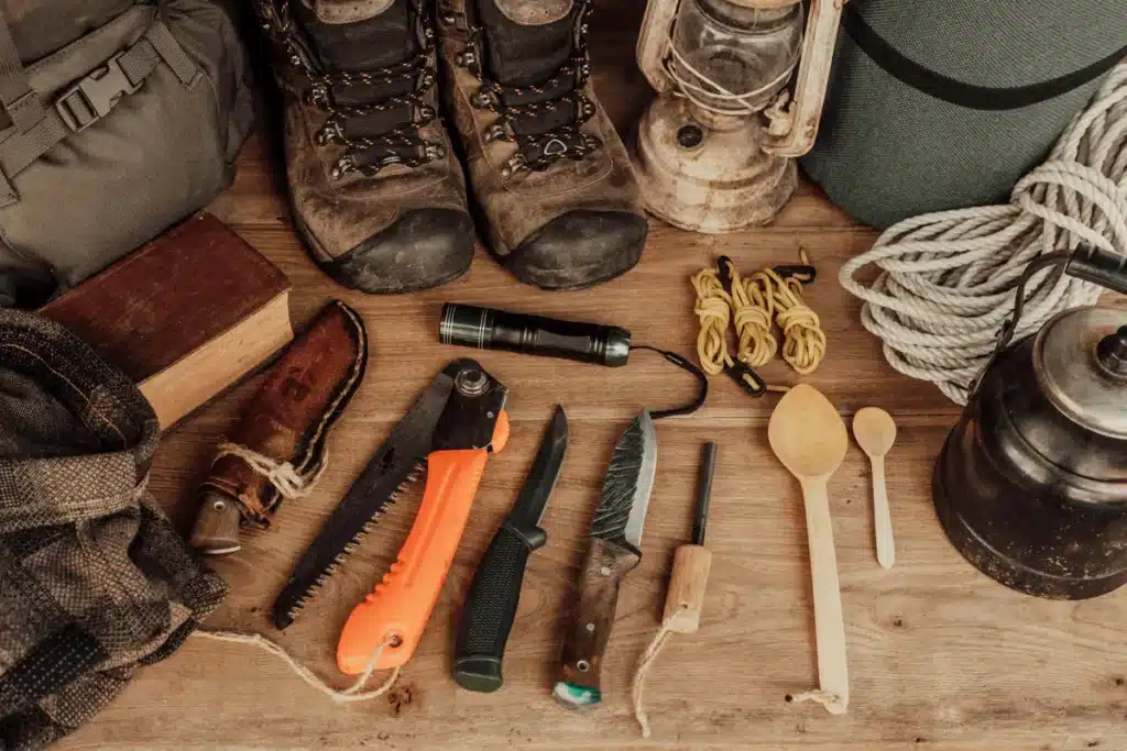 Uma visão de cima (flat lay) de um kit de Bushcraft sobre uma mesa de madeira. O kit inclui botas de trekking ou caminhada, uma serra dobrável laranja, diversas facas de sobrevivência (incluindo uma kukri), um lampião a querosene, um cantil ou chaleira escura, cordas, uma lanterna tática e utensílios de madeira. Os itens representam o equipamento essencial para Bushcraft e atividades de longa permanência na natureza.