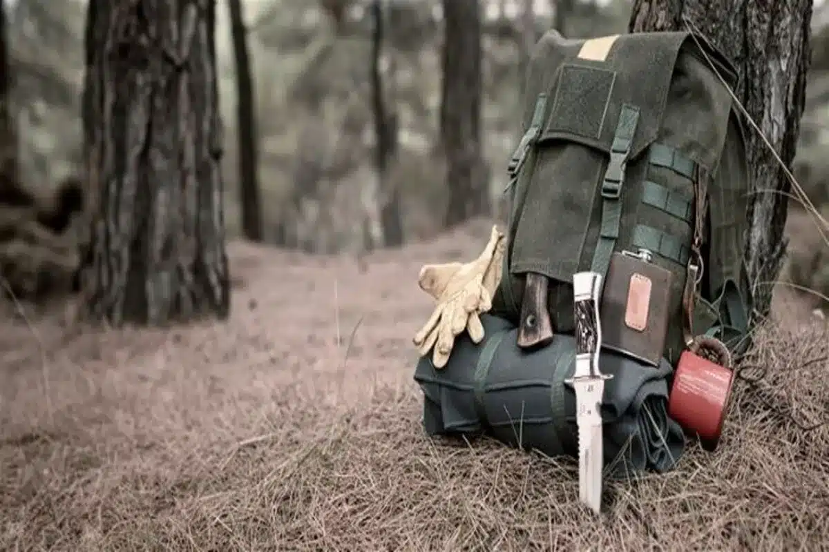 Uma mochila de lona verde de estilo militar, apoiada em uma árvore em uma floresta, com equipamentos de Bushcraft visíveis. Os itens incluem um facão ou faca de sobrevivência de lâmina fixa, luvas de couro, uma pochete de couro e uma caneca vermelha. A imagem, focada nos equipamentos em um chão coberto de agulhas de pinheiro, representa a preparação para a sobrevivência e acampamento na natureza selvagem.