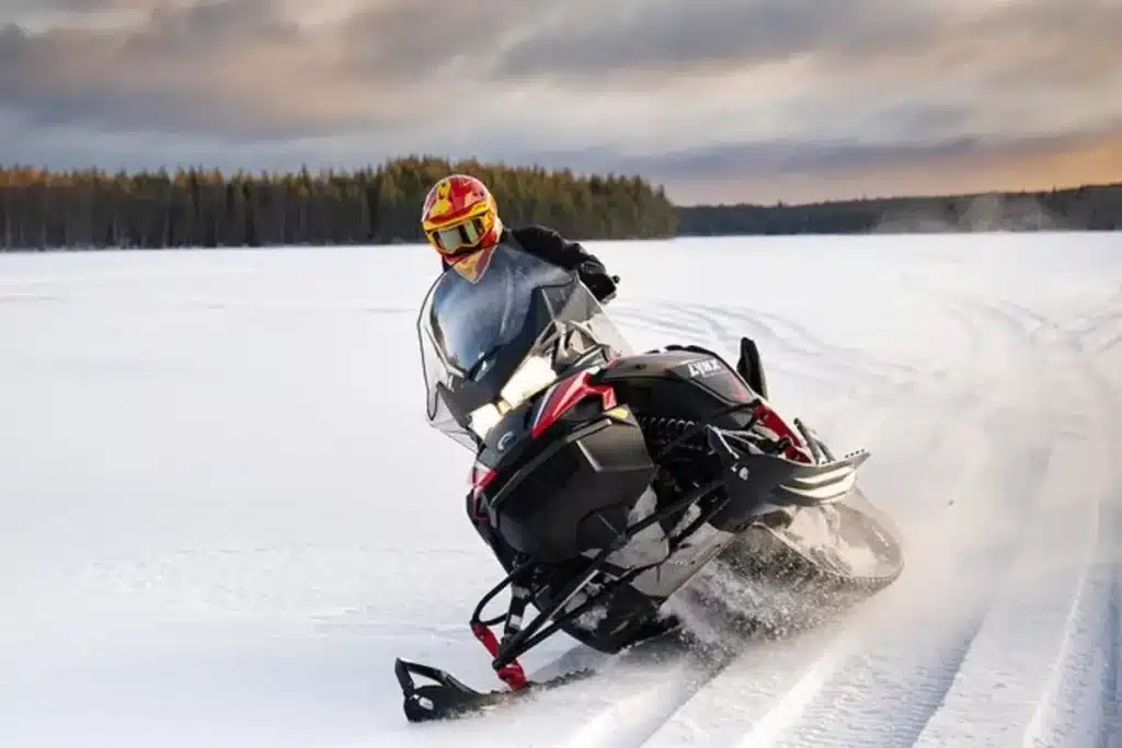 Um piloto usando capacete e óculos vermelhos e amarelos, fazendo uma curva acentuada com um Snowmobile (moto de neve) preto e vermelho, levantando neve em um campo aberto e congelado. A cena de ação ocorre em uma paisagem ártica ou nórdica, com uma floresta escura e um céu nublado ao fundo.