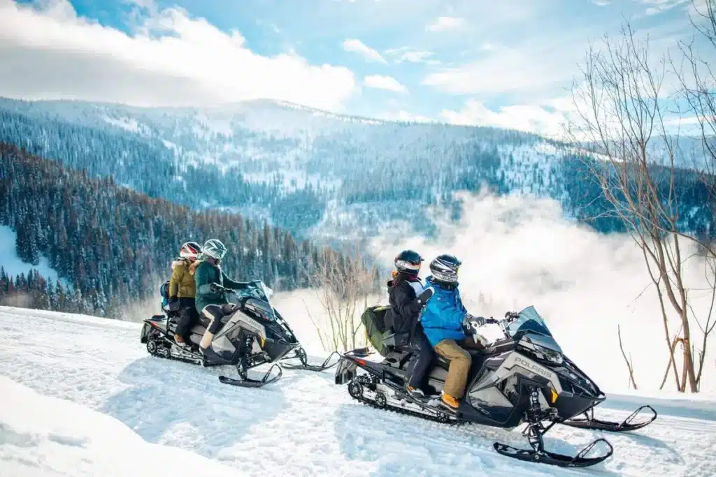 Duas duplas de pessoas pilotando Snowmobiles (motos de neve) em uma trilha nevada na montanha. Os pilotos estão usando capacetes de segurança e roupas de inverno em um dia ensolarado, com uma paisagem de montanhas cobertas por florestas e neve ao fundo, ideal para turismo de aventura.