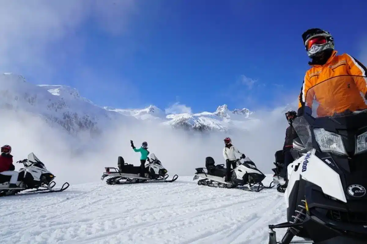Grupo de pessoas andando de snowmobile em uma área de montanha coberta de neve, com céu azul e picos nevados ao fundo, transmitindo aventura e diversão ao ar livre.