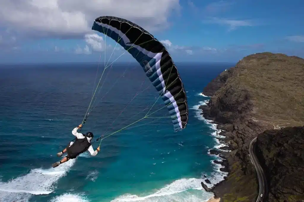Pessoa praticando Speed Flying com paraquedas preto e branco sobre mar azul e costa rochosa, em um céu parcialmente nublado.