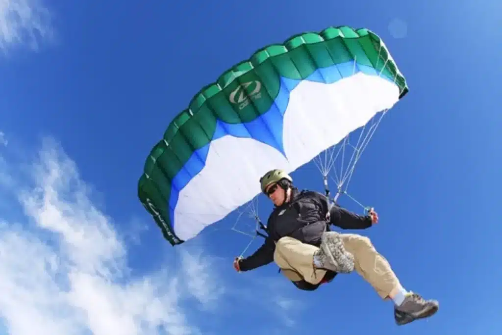 Pessoa praticando Speed Flying, voando próximo ao solo com um paraquedas esportivo verde e branco em um dia de céu azul.