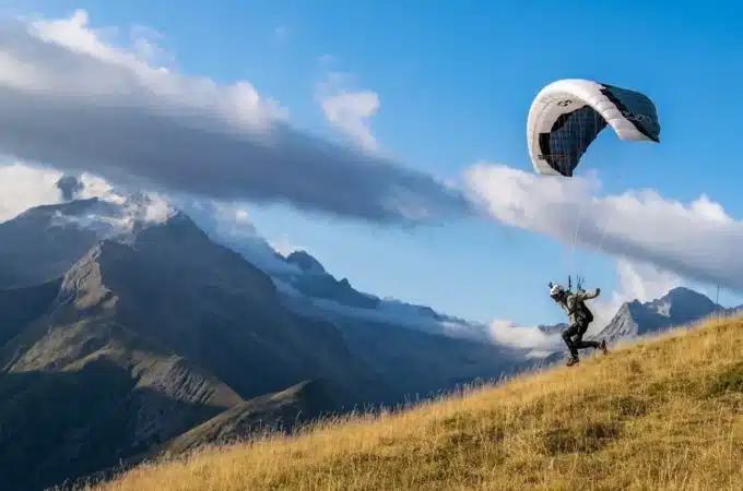Um homem pratica speed flying, correndo por uma encosta gramada de montanha com um parapente aberto acima dele. Montanhas majestosas e um céu azul com nuvens brancas são visíveis ao fundo, transmitindo uma sensação de aventura e liberdade.