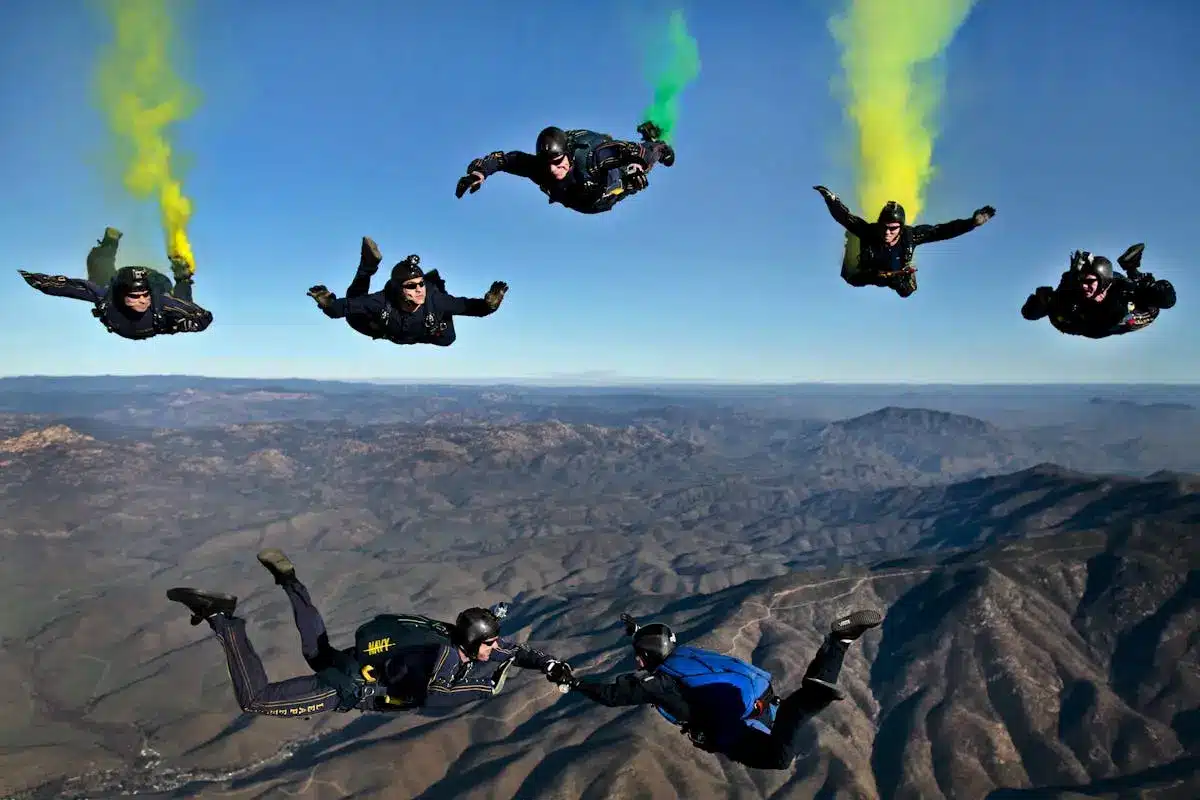 Grupo de paraquedistas realizando skydiving em formação sobre montanhas, com fumaça colorida e céu azul ao fundo