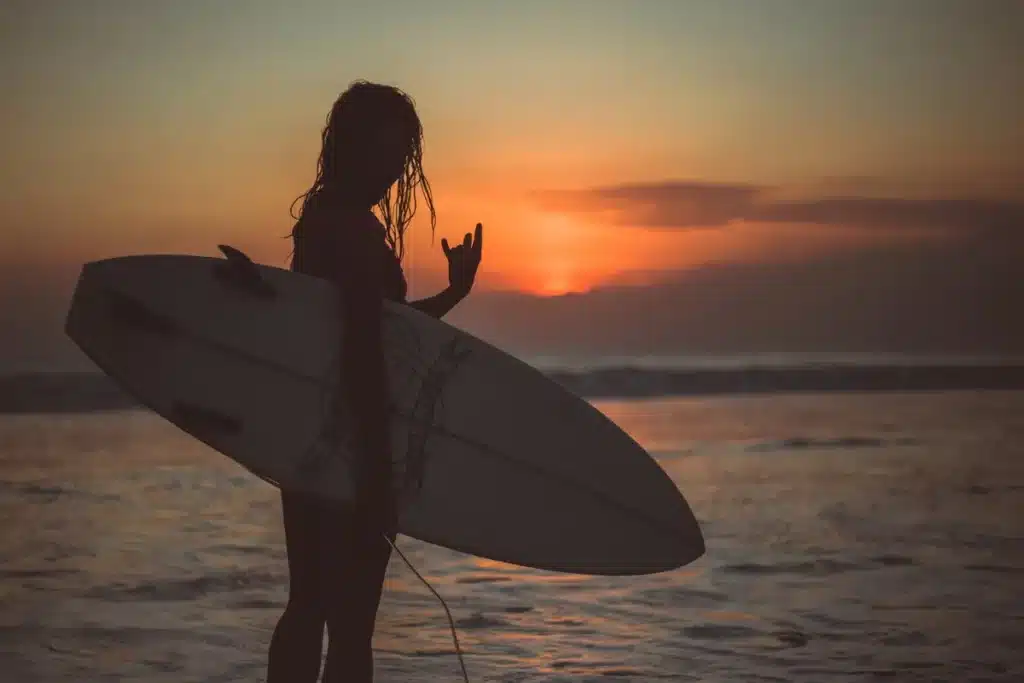 Silhueta de uma mulher surfista segurando uma prancha sob um pôr do sol alaranjado na praia, fazendo o sinal de shaka com a mão, transmitindo uma vibração de lazer e esporte radical.