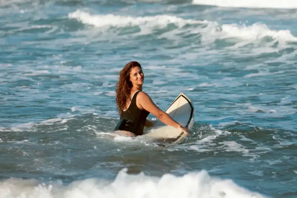 Uma mulher sorridente com cabelos cacheados entra no mar segurando uma prancha de surfe branca, com ondas quebrando ao seu redor, em uma cena de esporte aquático e lazer na praia.