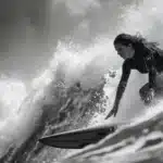 Foto em preto e branco de uma surfista em macacão de mergulho pegando uma grande onda, com foco na sua concentração e na força da água, ideal para imagens de esportes radicais e empoderamento feminino.