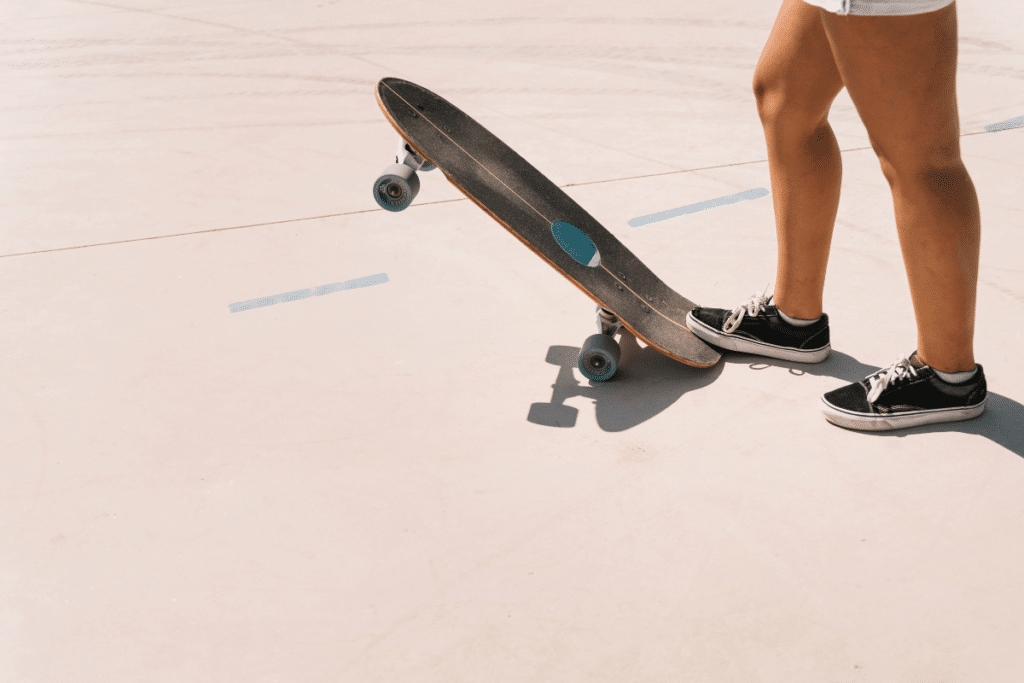 Vista lateral de uma mulher em shorts brancos e tênis pretos de skate, posicionada ao lado de um skate longboard escuro com estampa oval azul. O skate está inclinado sobre a roda de trás em uma pista de concreto claro sob sol forte.