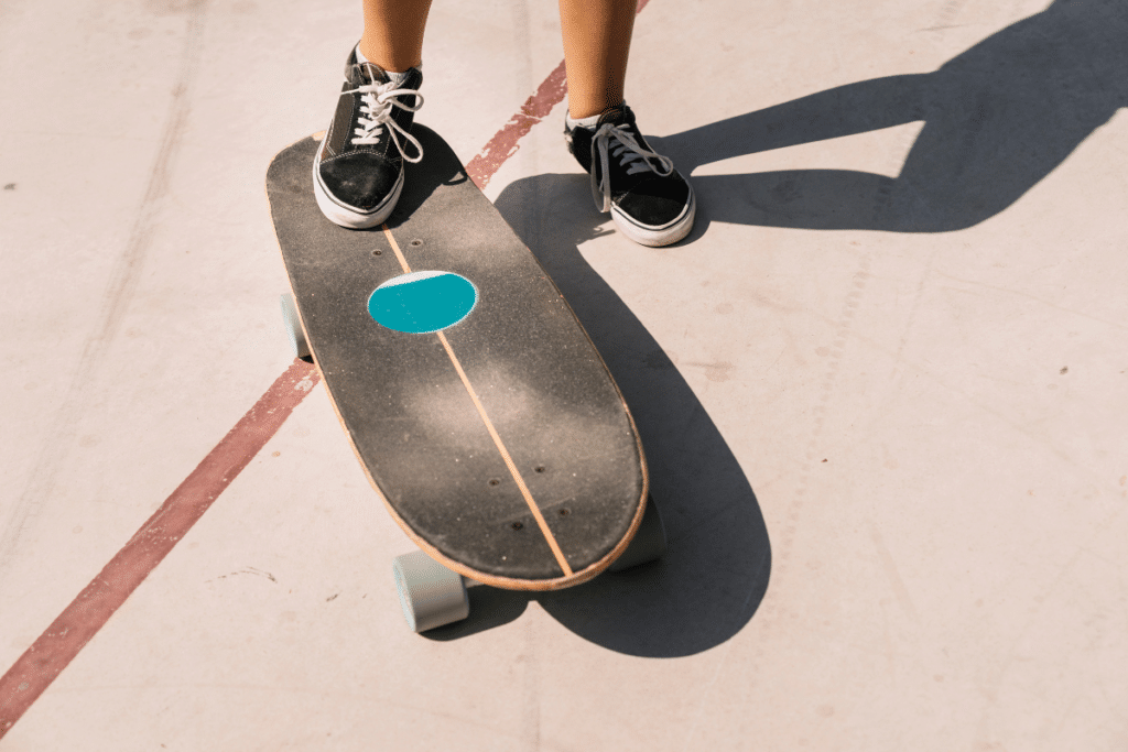 Vista de cima dos pés de uma pessoa em tênis pretos de lona com cadarços brancos, posicionados em um skate longboard ou cruiser escuro e desgastado, com um decalque oval azul-turquesa central. O skate está sobre uma superfície de concreto claro, com uma linha vermelha e sombra forte do skatista.