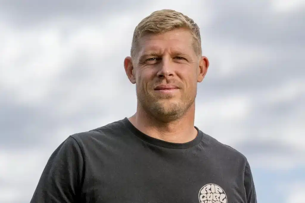 homem de camiseta preta olhando para a câmera em um ambiente externo com fundo desfocado