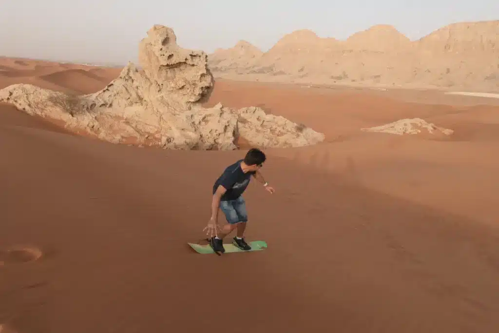 pessoa andando de sandboard em duna em área desértica com formações rochosas ao fundo