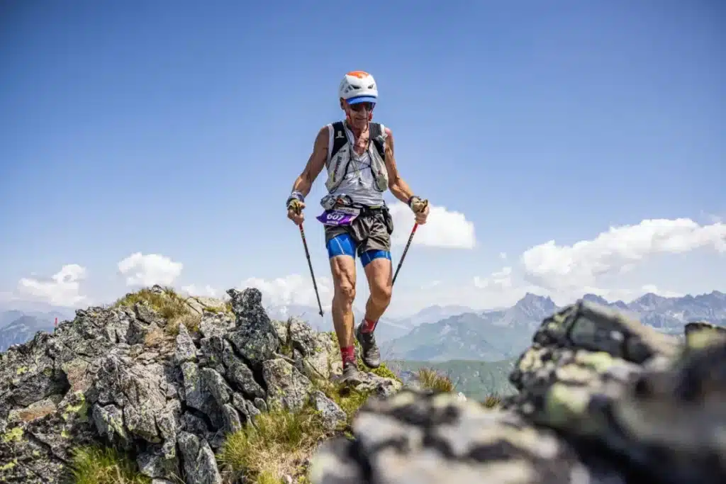 Atleta de corrida em montanha caminhando sobre pedras no topo de uma montanha usando bastões de apoio.