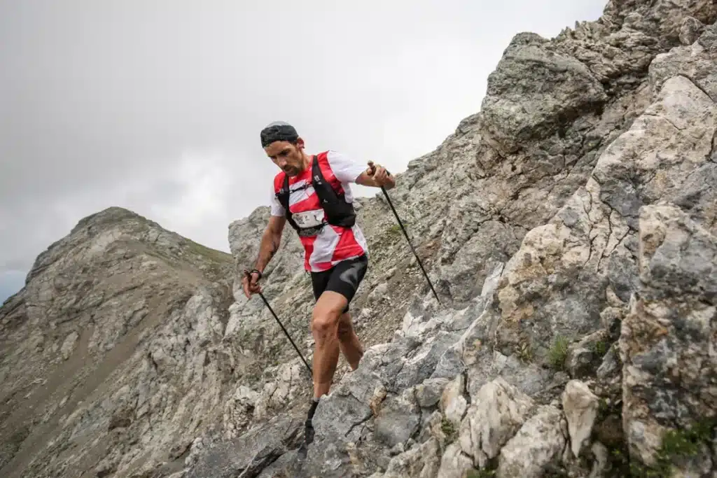 Corredor descendo uma trilha íngreme e rochosa em ambiente de montanha usando bastões de trekking.