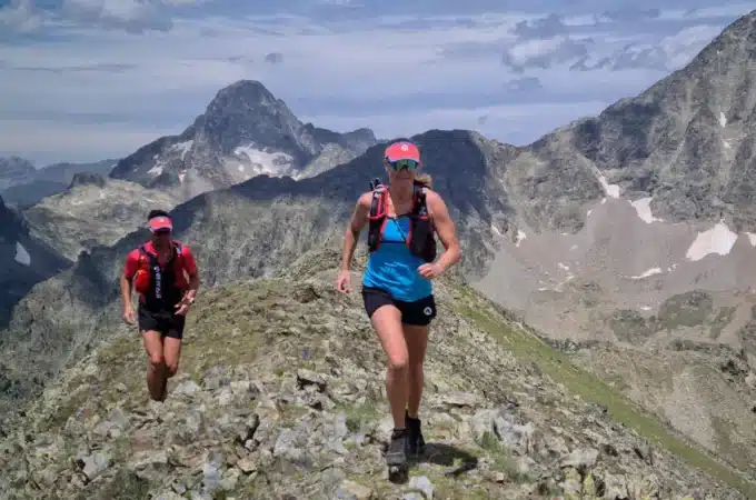 Dois corredores praticando skyrunning em uma trilha estreita no topo de uma montanha rochosa com picos ao fundo.