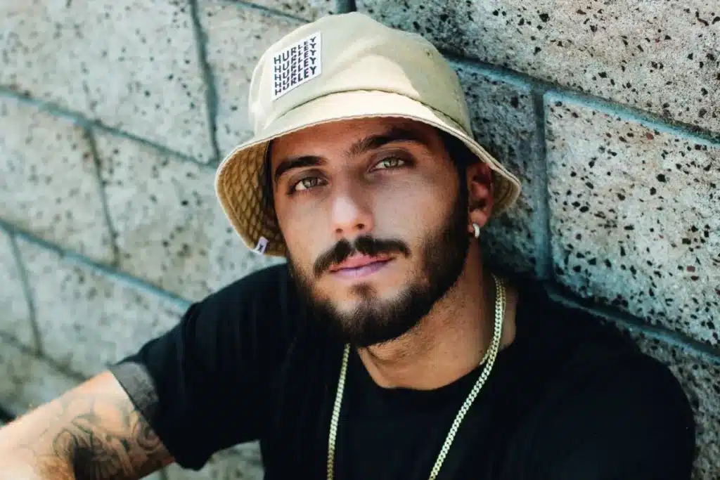 homem com chapéu bucket e camiseta preta encostado em parede de concreto olhando para a câmera