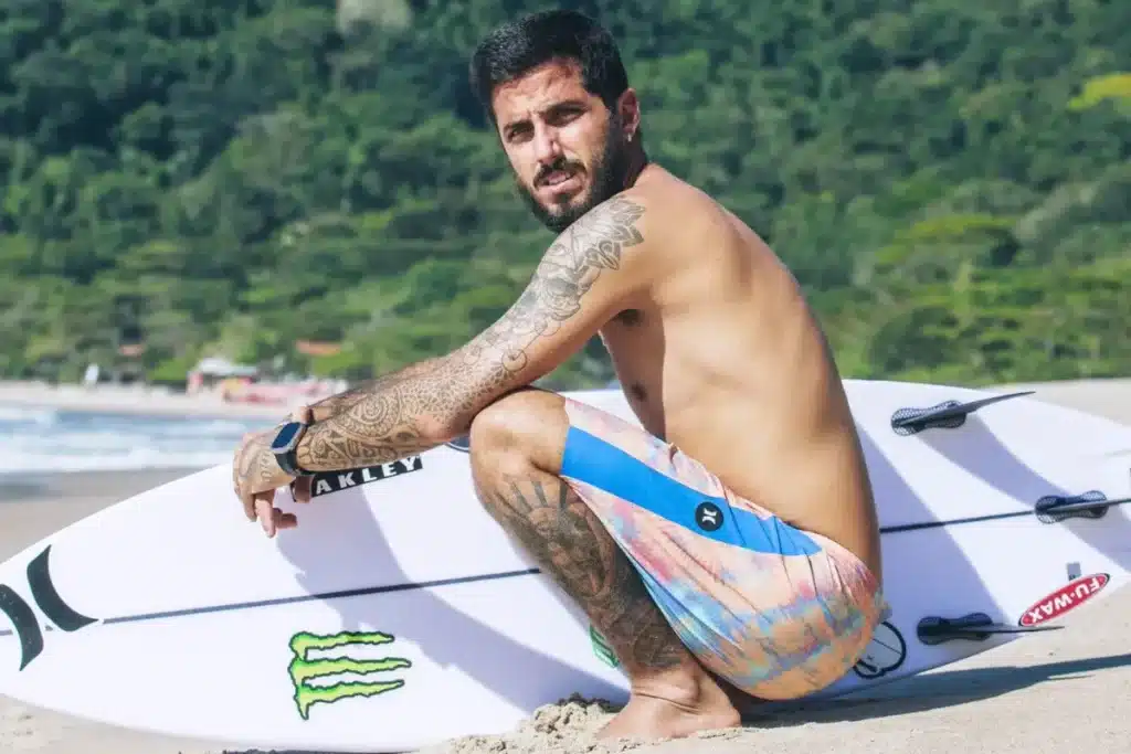 surfista sentado na areia segurando prancha de surf branca em uma praia com vegetação ao fundo
