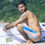 surfista sentado na areia segurando prancha de surf branca em uma praia com vegetação ao fundo