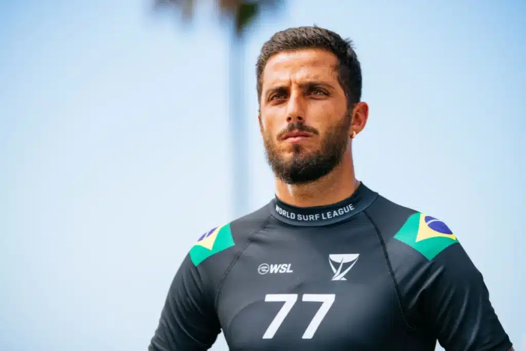 surfista usando lycra preta com número 77 durante competição de surf com bandeira do Brasil nos ombros