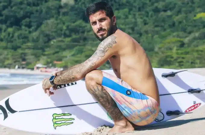 surfista sentado na areia segurando prancha de surf branca em uma praia com vegetação ao fundo