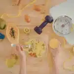 mesa com alimentos saudáveis, halteres, fita métrica e balança ao lado de salada e suco natural