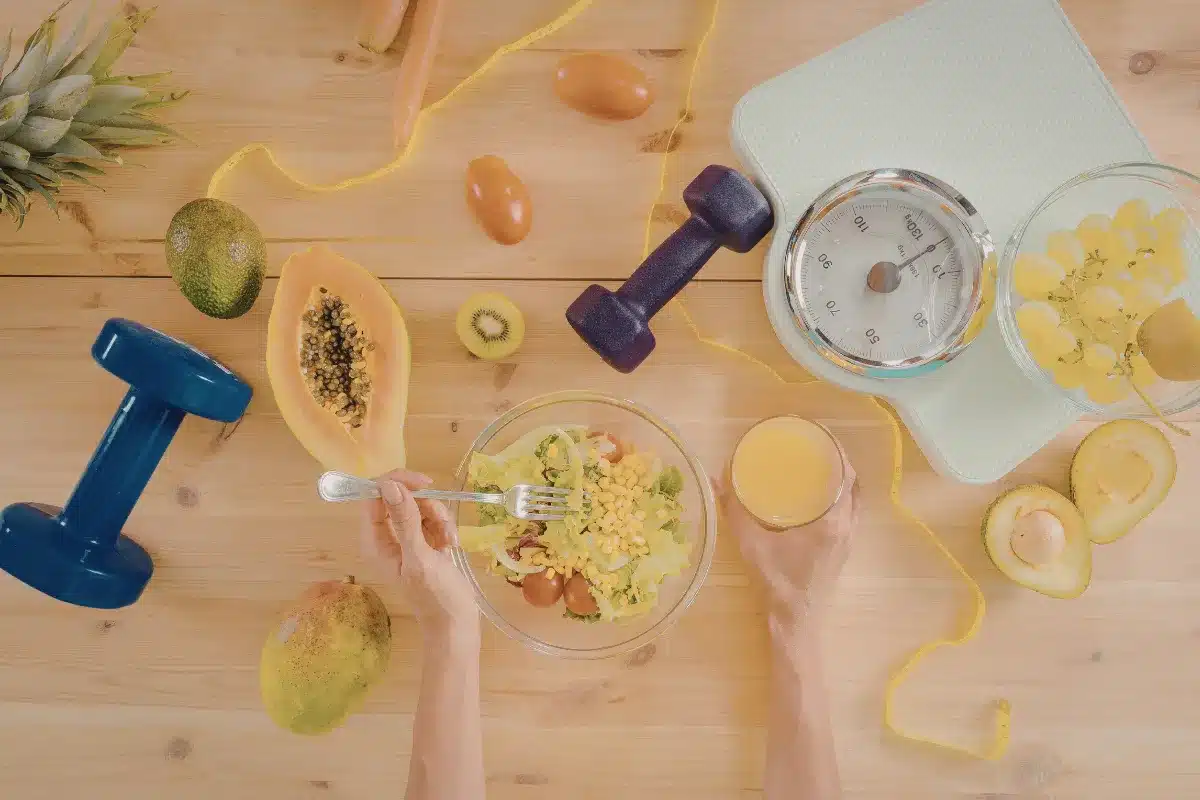 mesa com alimentos saudáveis, halteres, fita métrica e balança ao lado de salada e suco natural